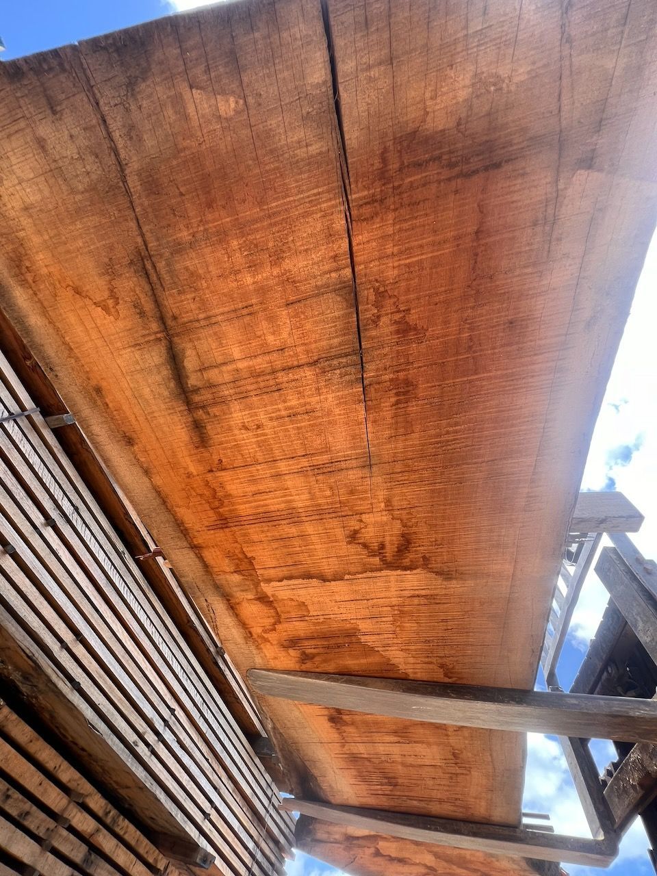 Wooden planks stacked, some with a visible grain, outdoors under a blue sky.