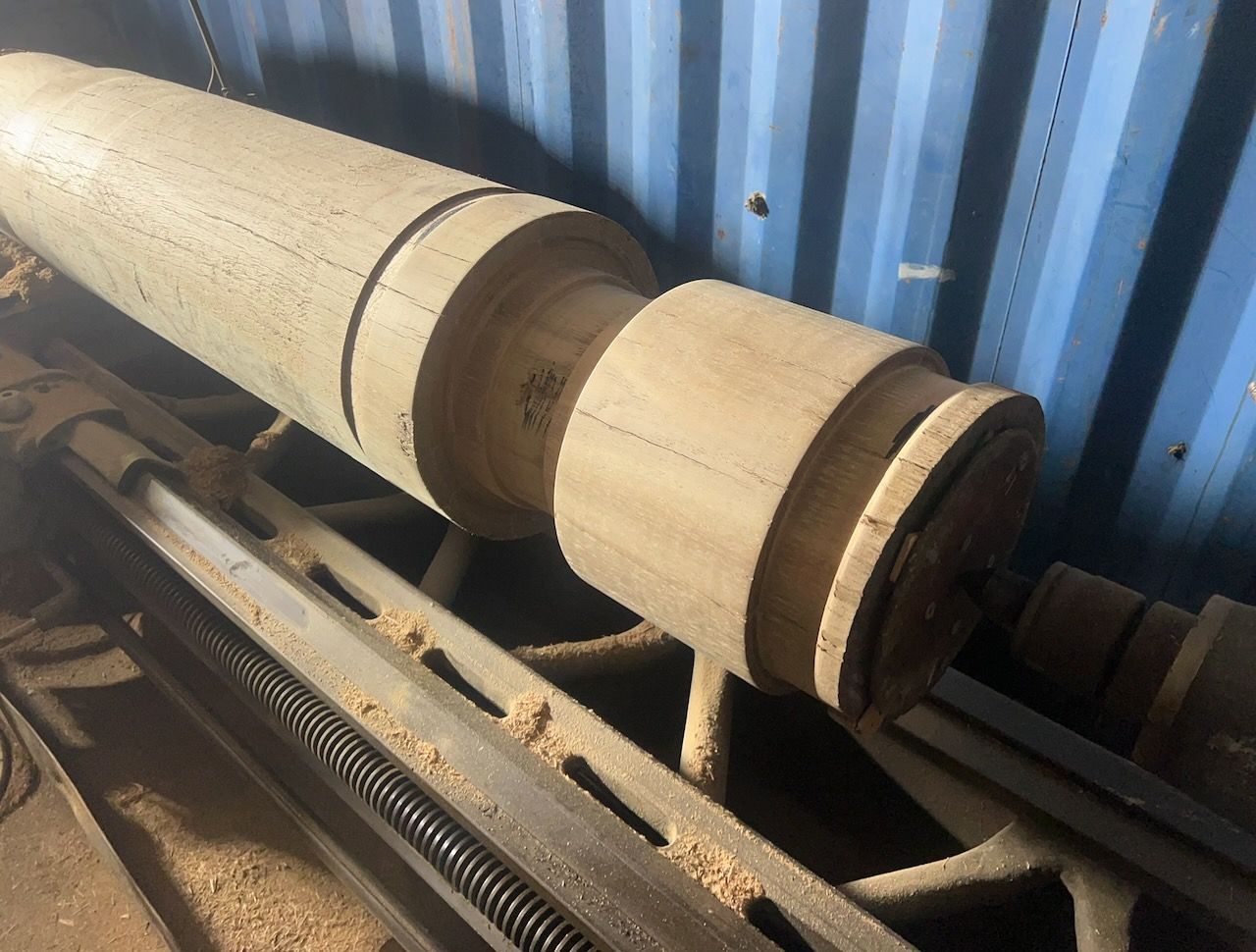 A large wooden spindle on a lathe, surrounded by wood shavings in a workshop.