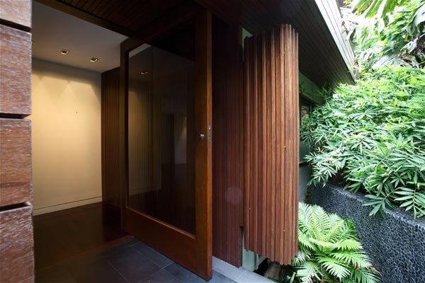 A modern entrance with a wooden pivot door, recessed lighting, and lush green plants in a stone planter.