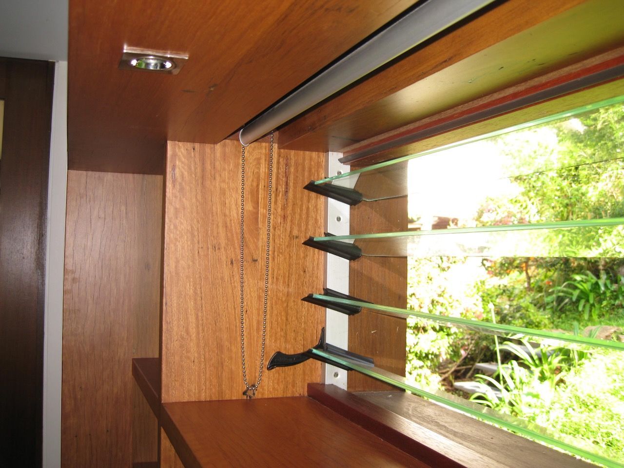 Wooden louvered window with open panes, chain to adjust angle, and view of greenery outside.