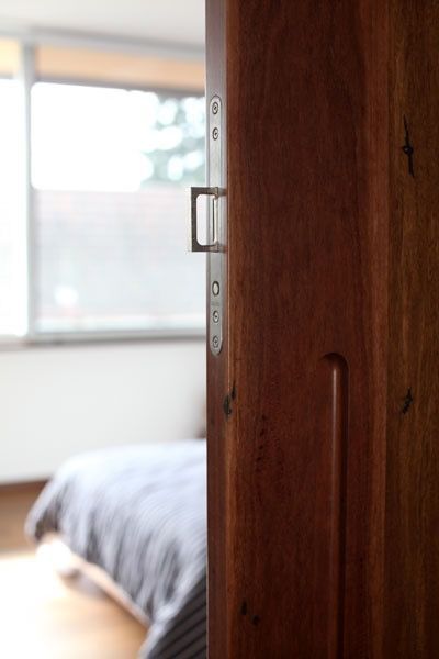Wooden door slightly ajar, revealing a bed and window in a bedroom. Silver door latch visible.