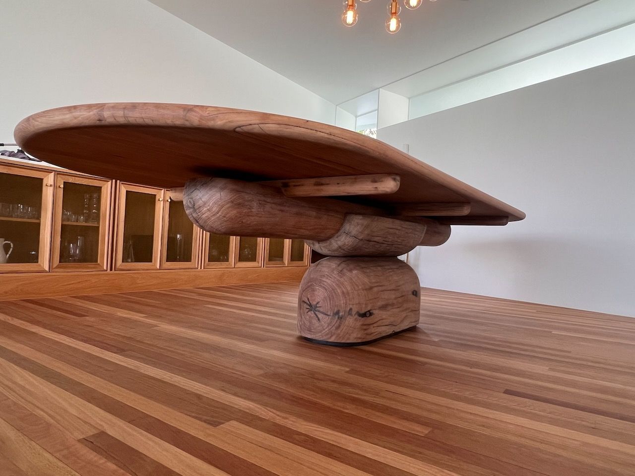 A large, oval wooden dining table with a thick, rustic log-style pedestal base sits on a matching hardwood floor.