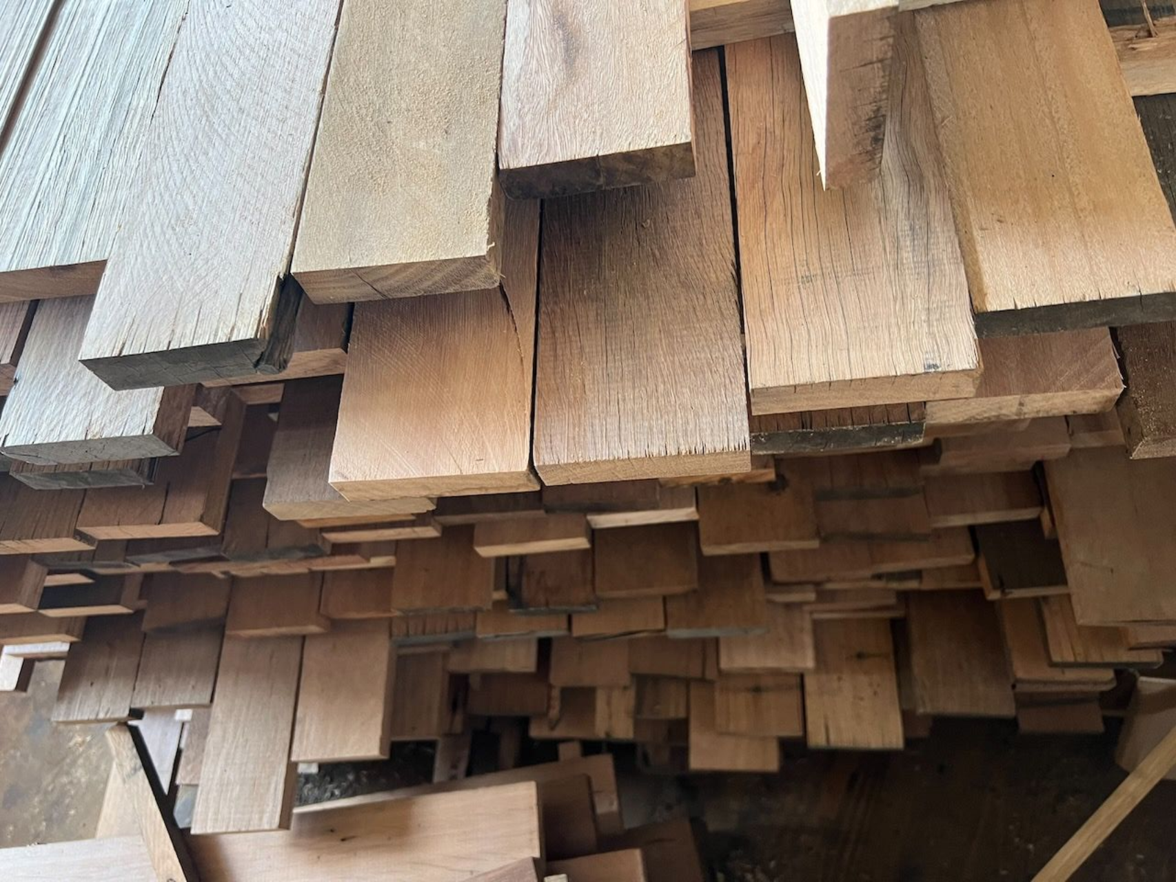 A large, dense stack of rough-cut, unfinished wooden boards piled neatly in a workshop or lumber yard.