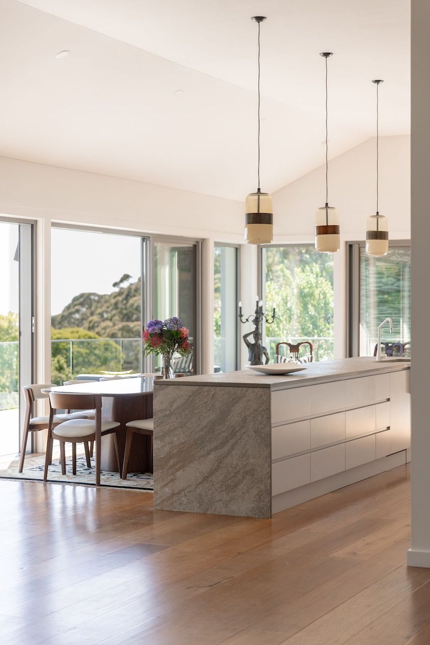 Modern kitchen with a large island, windows, and dining area overlooking a green landscape.
