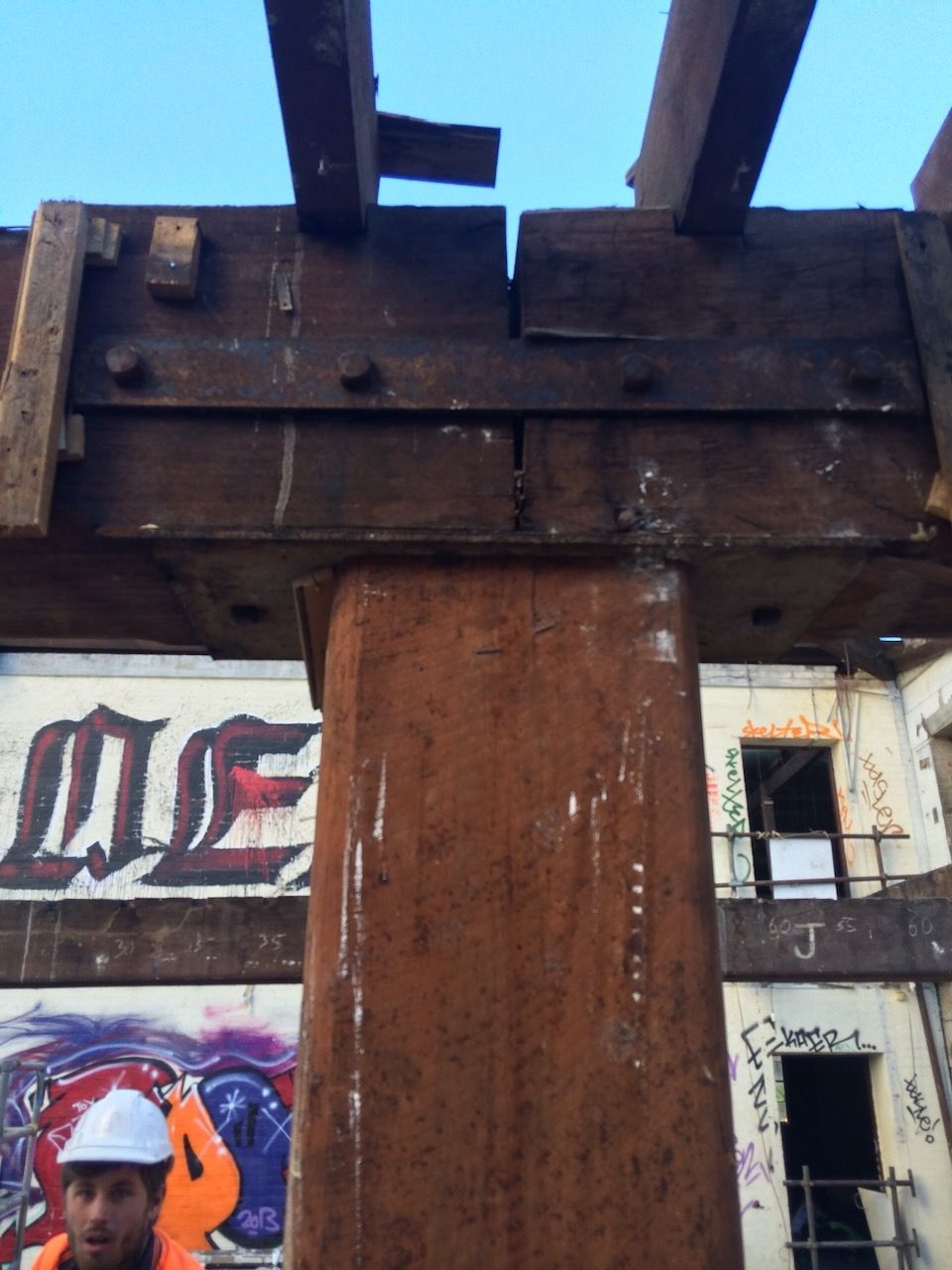 A construction worker in a white hard hat stands beneath a large timber beam joint supported by a central wood post.
