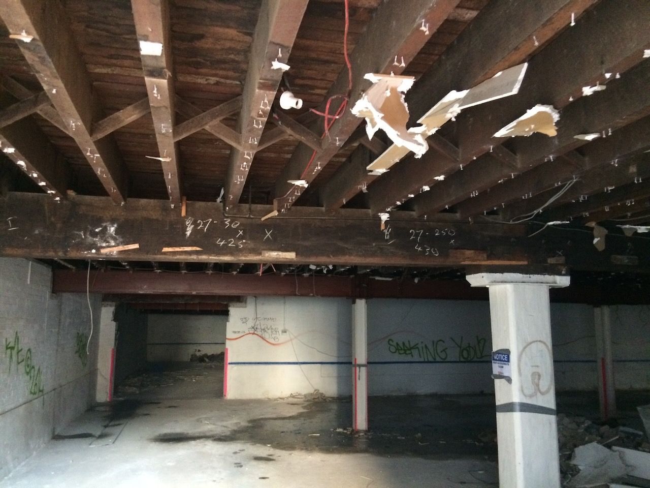 An unfinished interior space with exposed wooden ceiling joists, steel support beams, columns, and debris-strewn floors.