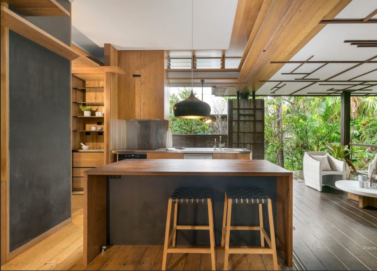 Modern kitchen with wooden cabinetry and island, overlooking a lush outdoor space with seating.