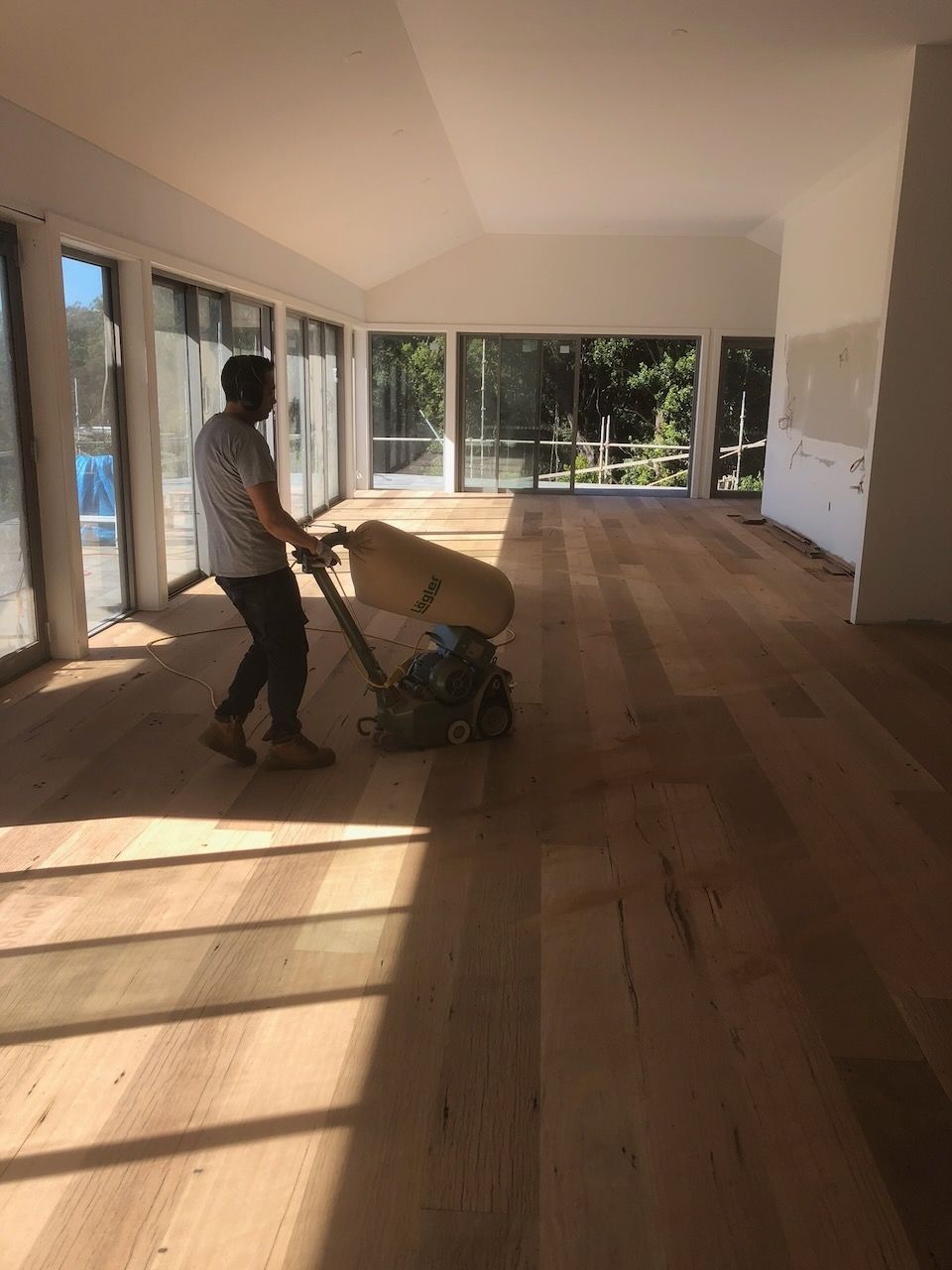 Person sanding a wood floor with a large machine in a sunlit room with large windows in Brisbane.