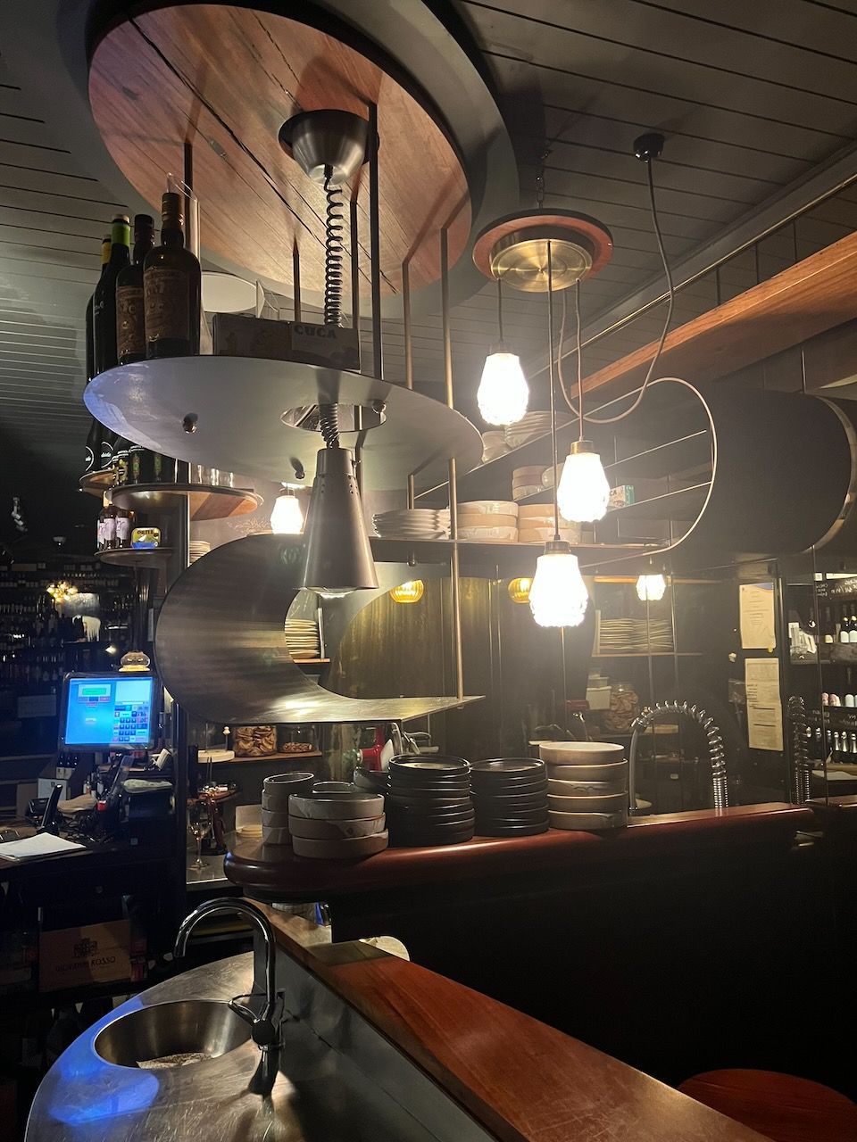 A restaurant interior with a spiral shelf and hanging lights above a bar with dishes.