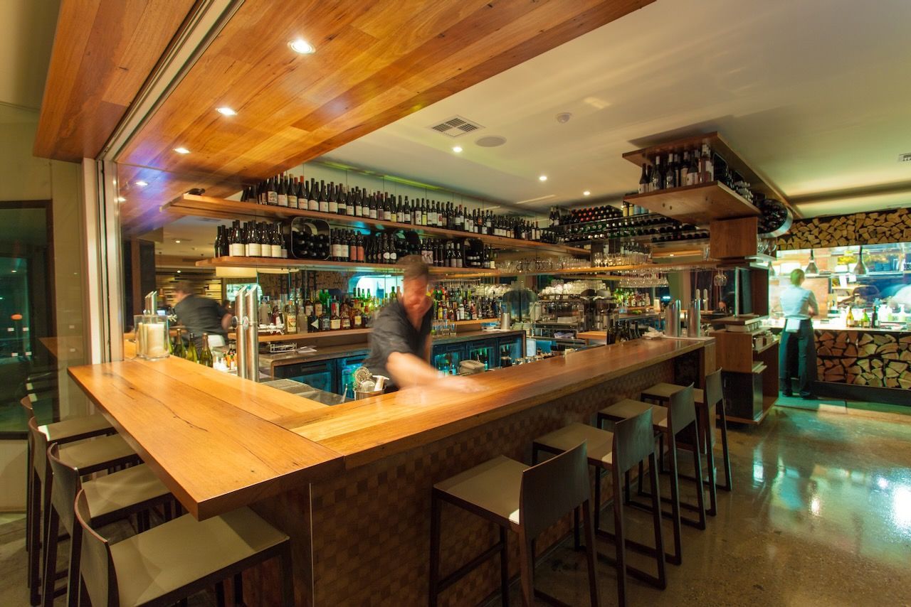 A modern bar with a wooden counter and stools, featuring shelves of bottles and warm lighting, with a worker in motion.