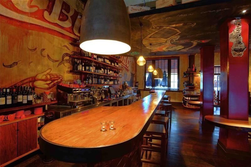 Interior view of a bar with a long wooden counter, liquor bottles, and hanging lights; mural on the wall.