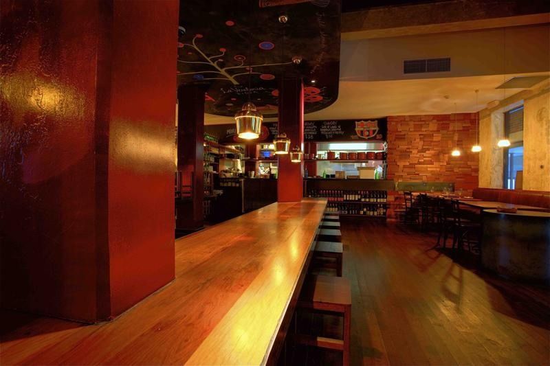 Long wooden bar in a dimly lit restaurant with red accents, bar stools, and tables.