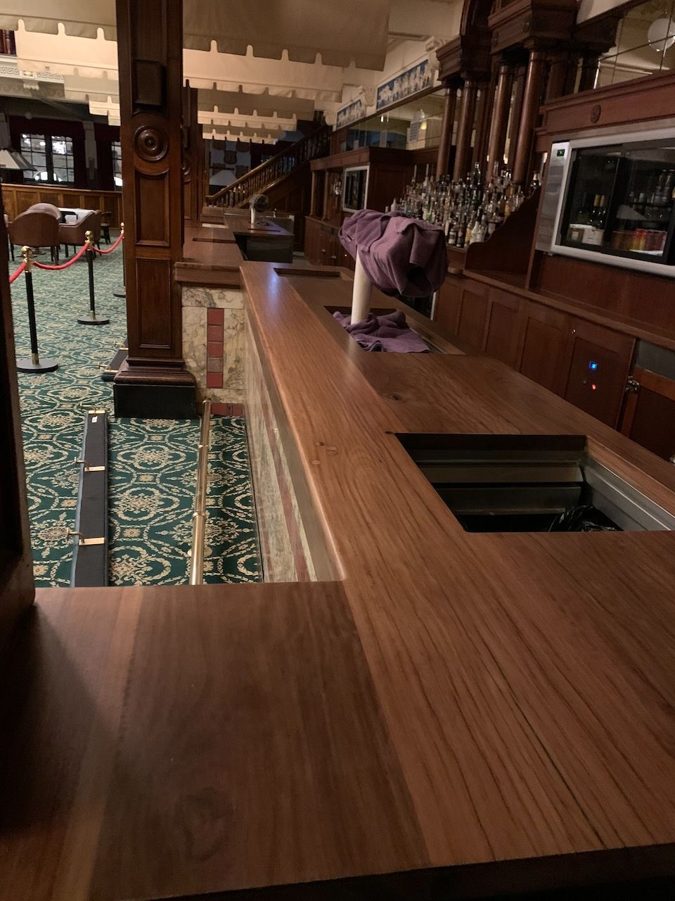 Wooden bar in a dimly lit room, with taps, a green patterned carpet, and a partially visible mirror.
