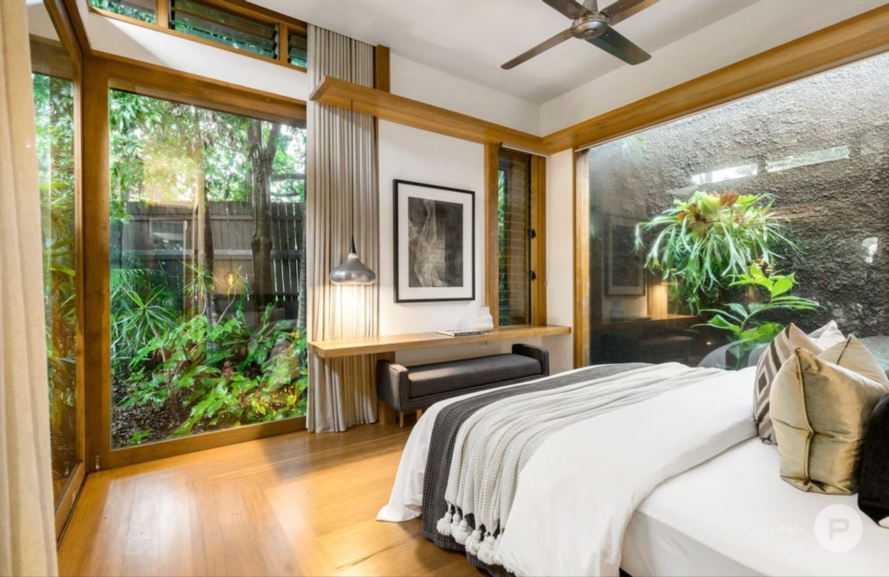 Modern bedroom with large windows overlooking lush greenery, a wooden desk, and a neatly made bed with neutral bedding.