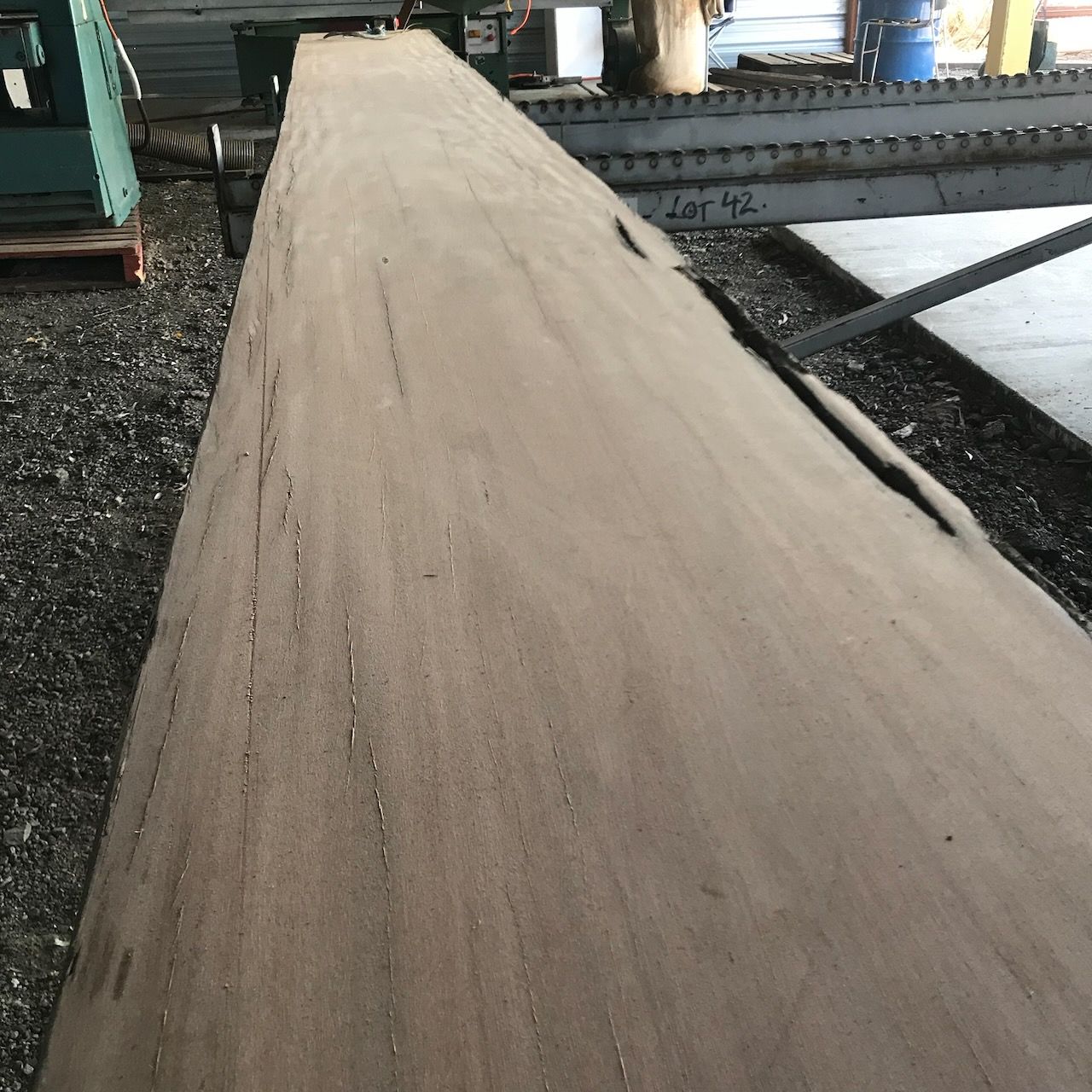 A long, light brown wooden plank on a rough surface, possibly at a lumberyard.