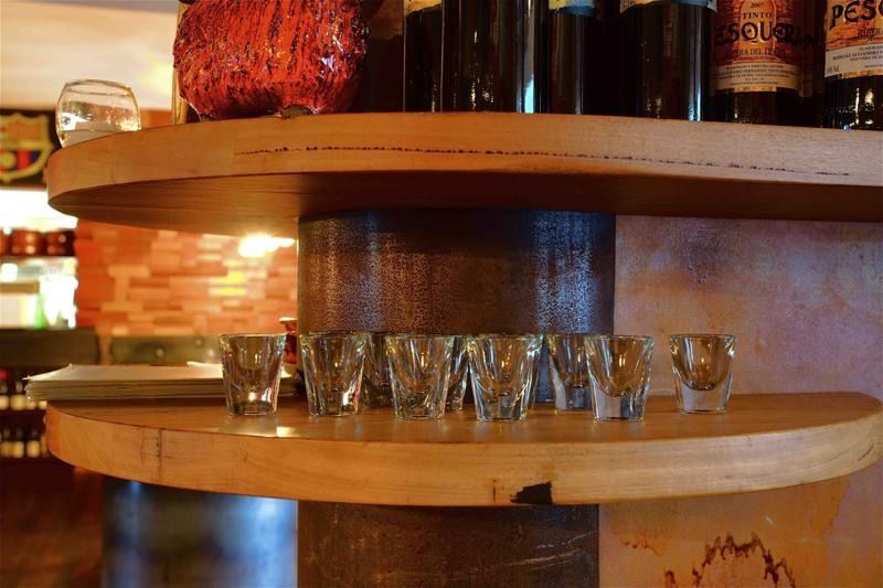 Shot glasses on a wooden shelf in front of bottles, with a brick wall visible in the background.