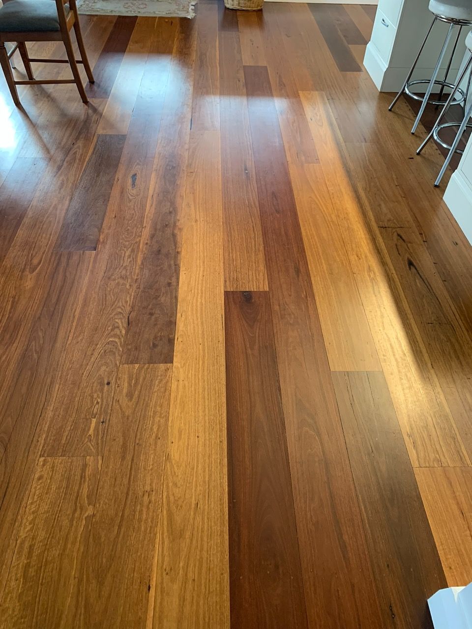 Hardwood flooring with varied brown, amber, and tan planks in a kitchen setting with bar stools and chair legs visible.
