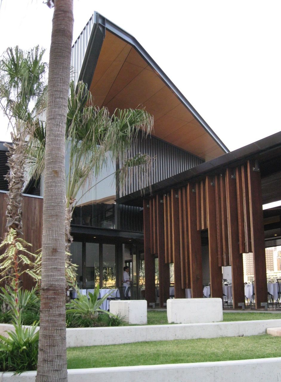 A modern building with a steep, angled roof and wood-slatted facade, set behind terraced grass and palm trees.