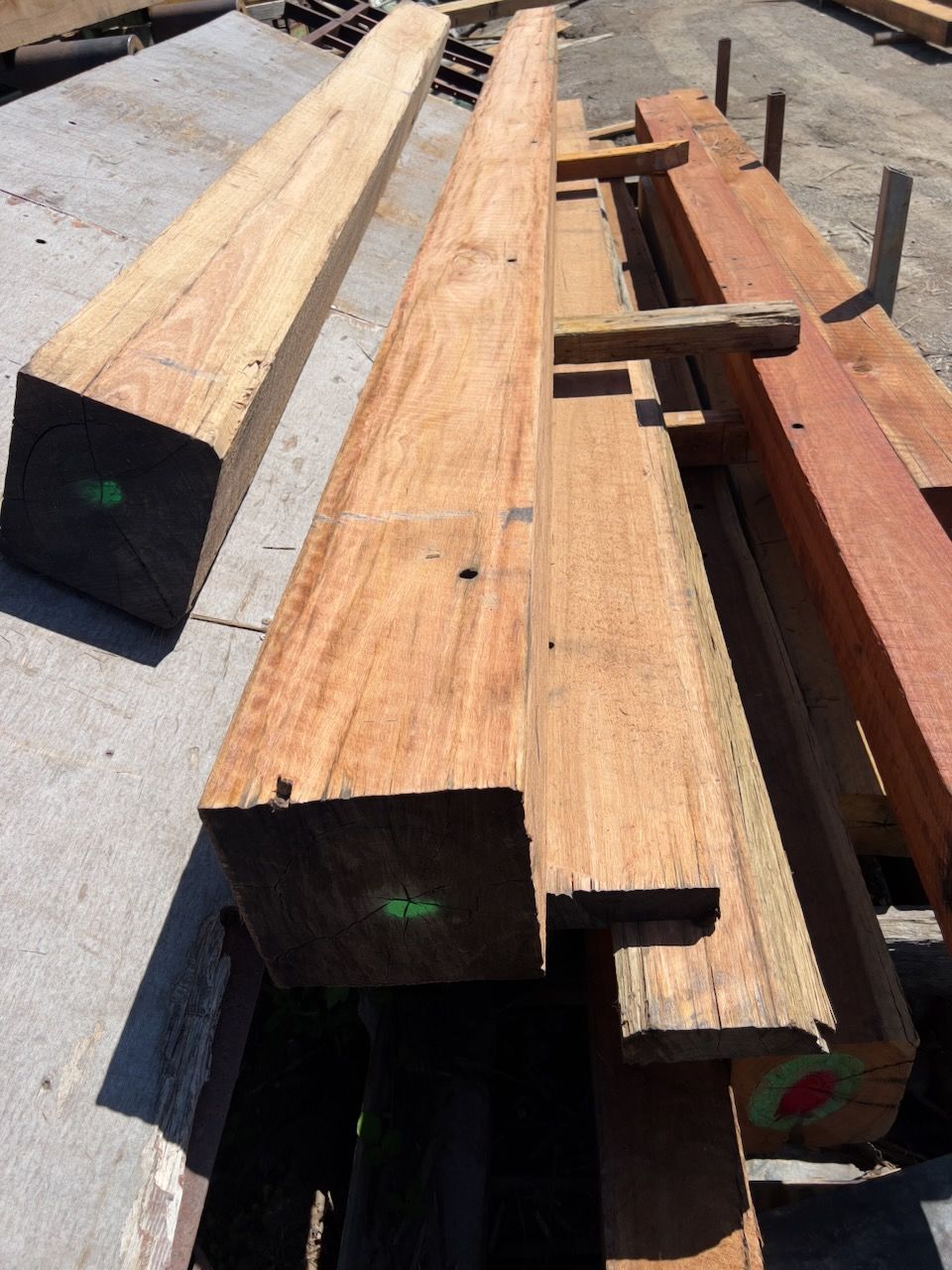 Wooden lumber stacked outdoors, varying sizes and reddish-brown color, with green and red markings.
