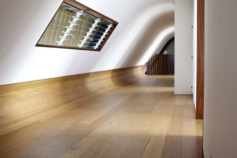 A modern hallway with light wood flooring, curved white walls, and a sloped skylight, leading toward a wooden staircase.