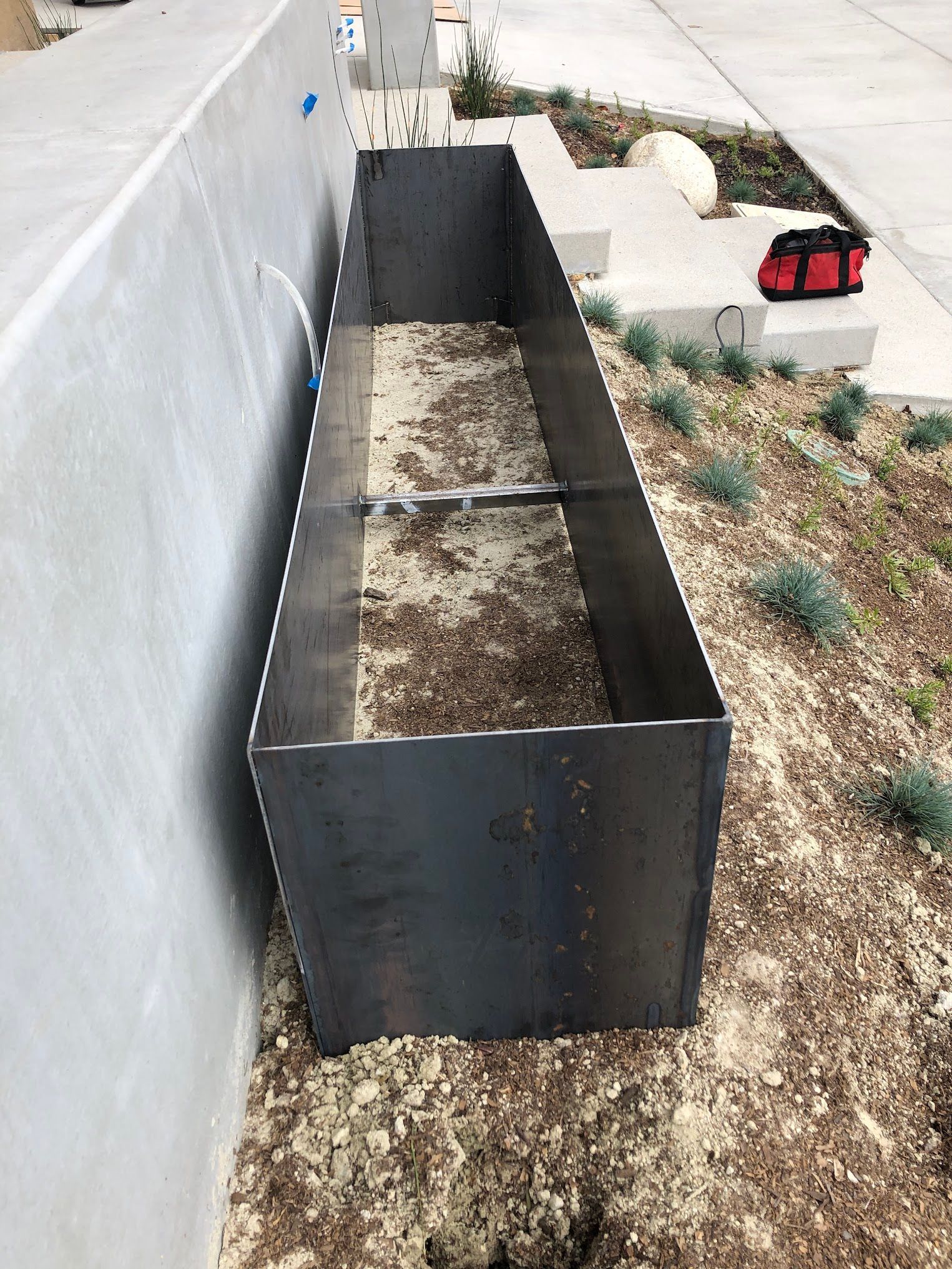 A metal planter is sitting in the dirt next to a wall.