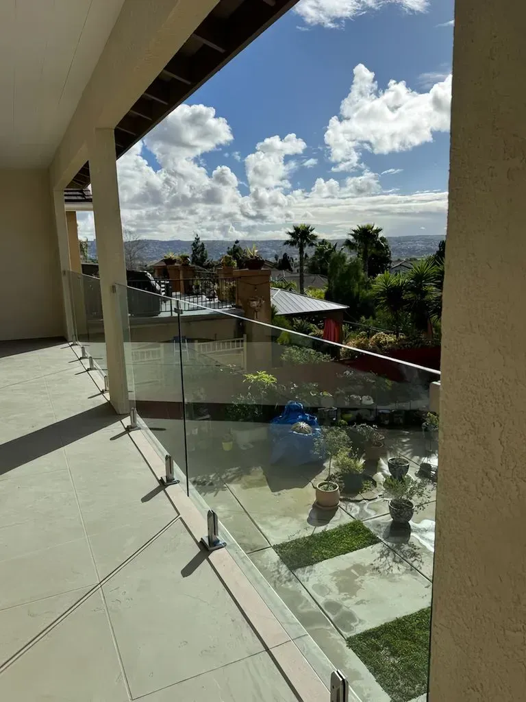 Balcony view of a city, featuring glass railing, blue sky, and a lush garden.
