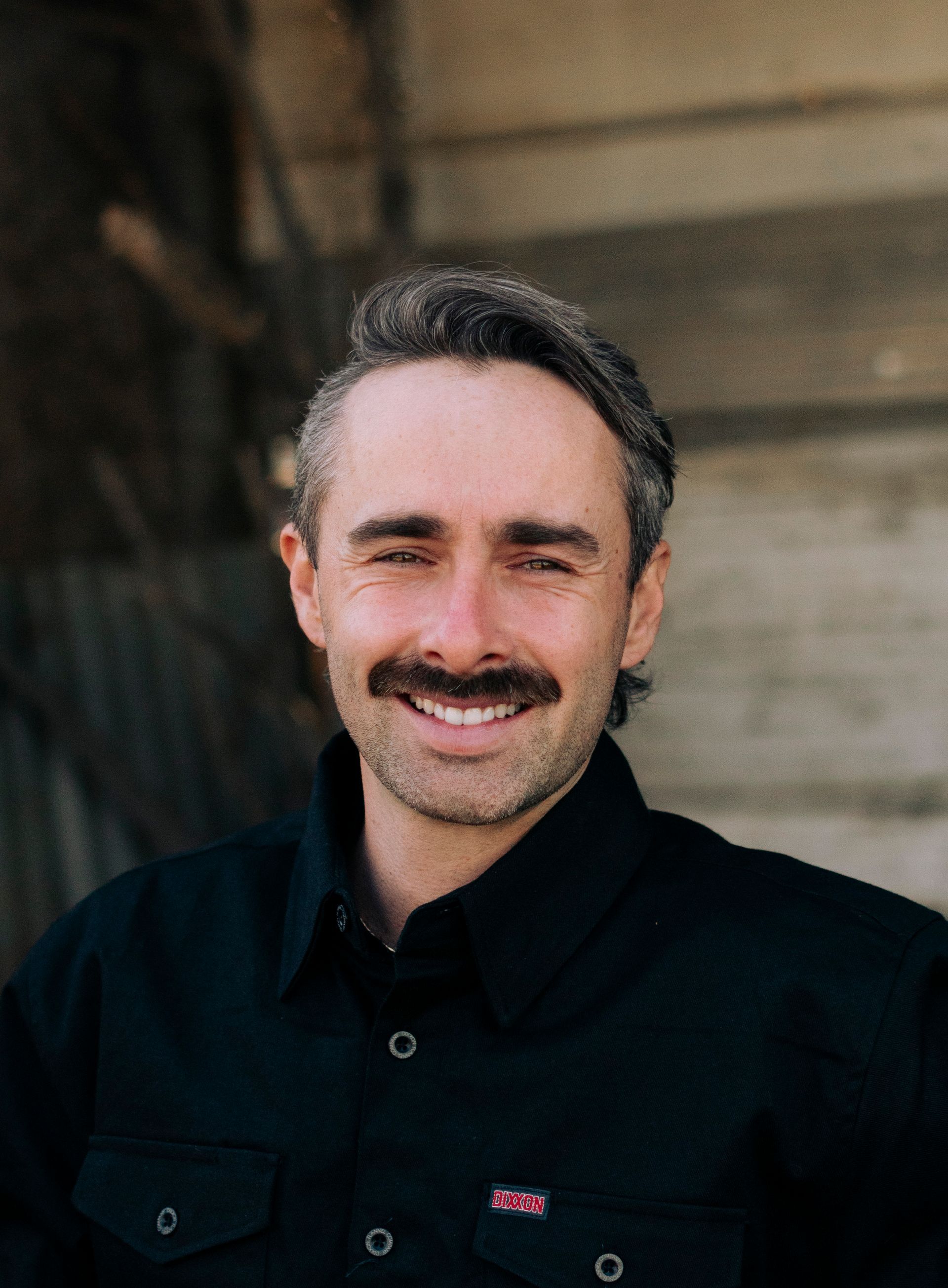 A man in a suit and black shirt is standing in front of a brick wall.