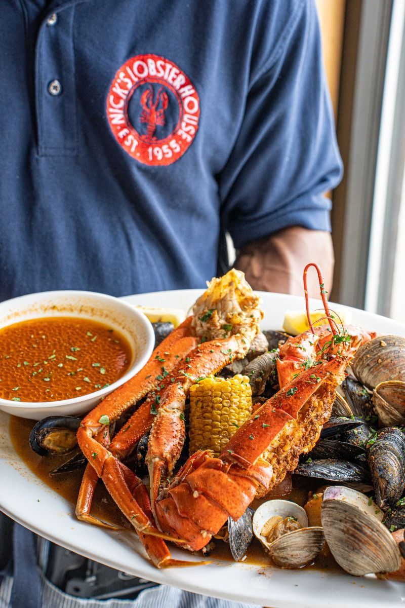 Person holding a platter of seafood: lobster, crab legs, corn, mussels, clams, and dipping sauce.