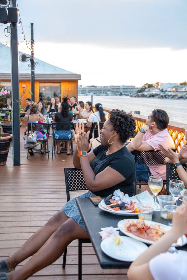 Woman clapping at waterfront restaurant; others watch. Cloudy sky, water view.