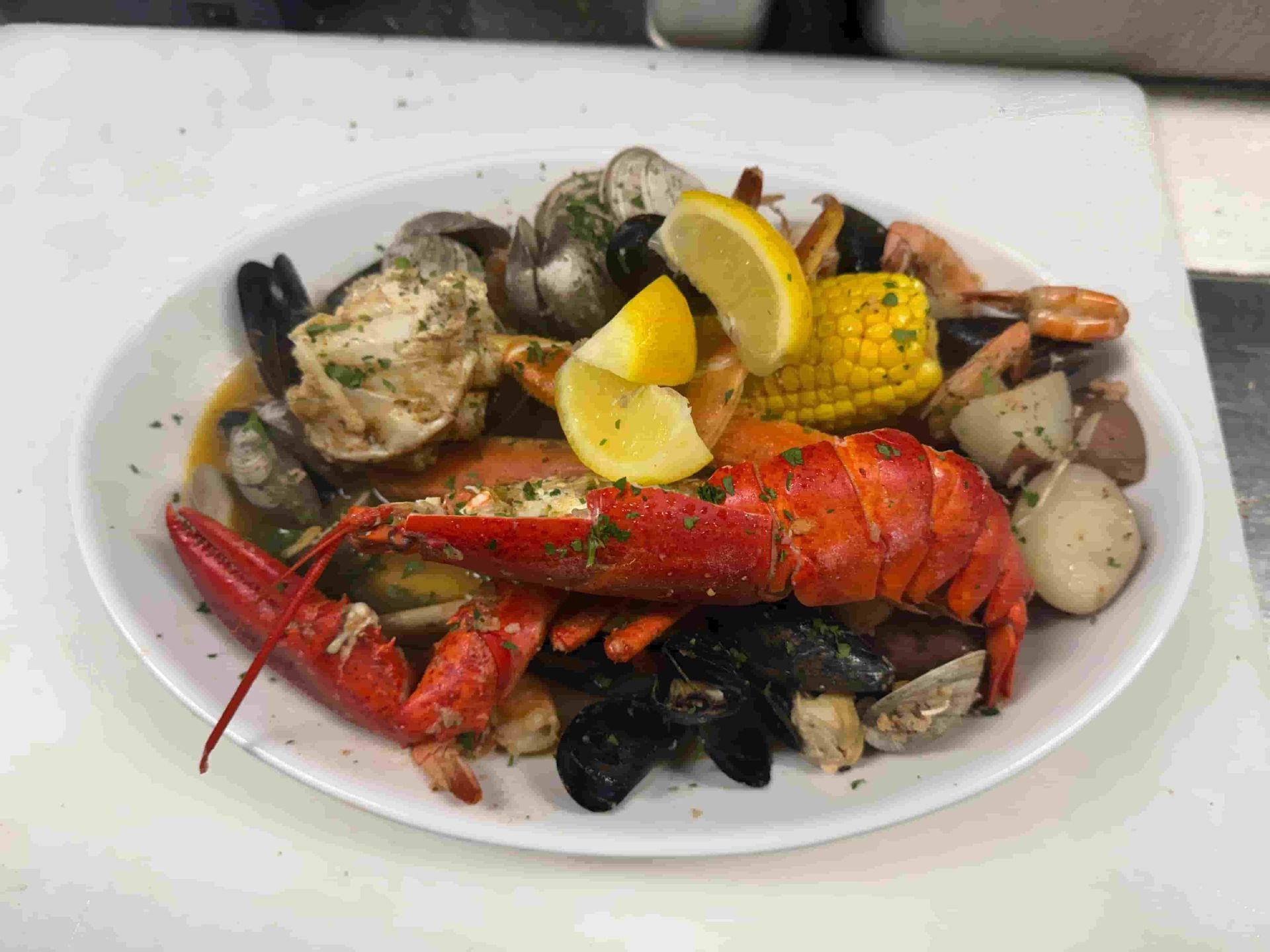 Seafood boil on a white plate: lobster, clams, mussels, shrimp, corn, potatoes, lemon slices.