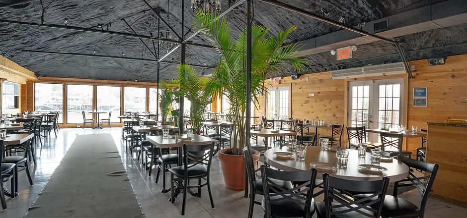 Restaurant interior with tables, chairs, and plants; wood paneling, large windows, and a dark ceiling.