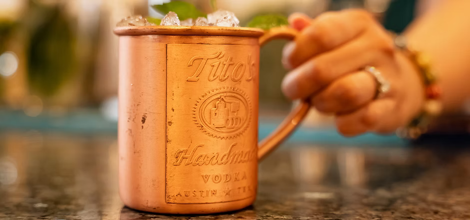 A copper mug of cocktail, with crushed ice and a mint garnish, is held by a hand with a ring.