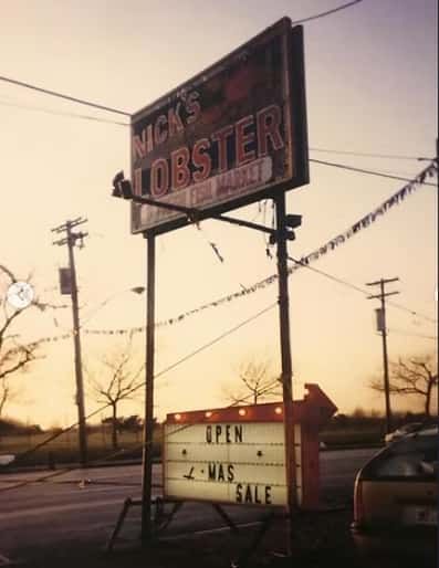Sign for Mick's Lobster Fish Market. Open sign below. Roadside with power lines and trees at dusk.
