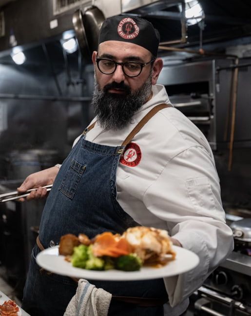 Chef in white coat and denim apron presents a plate with food in kitchen.