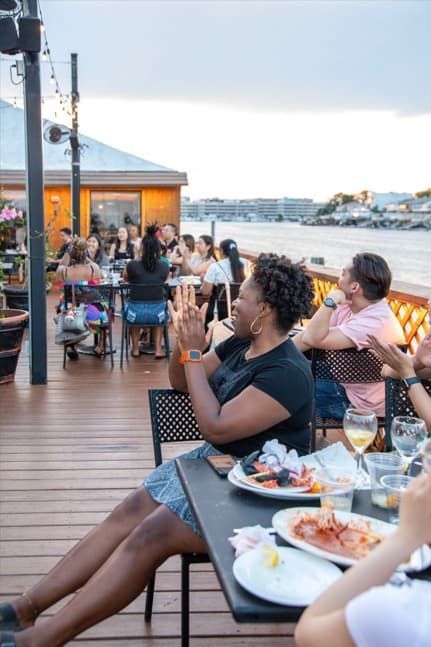 Woman clapping at waterfront restaurant; others watch. Cloudy sky, water view.