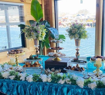 Dessert table set for an event; turquoise tablecloth with treats, flowers, by a waterfront window.