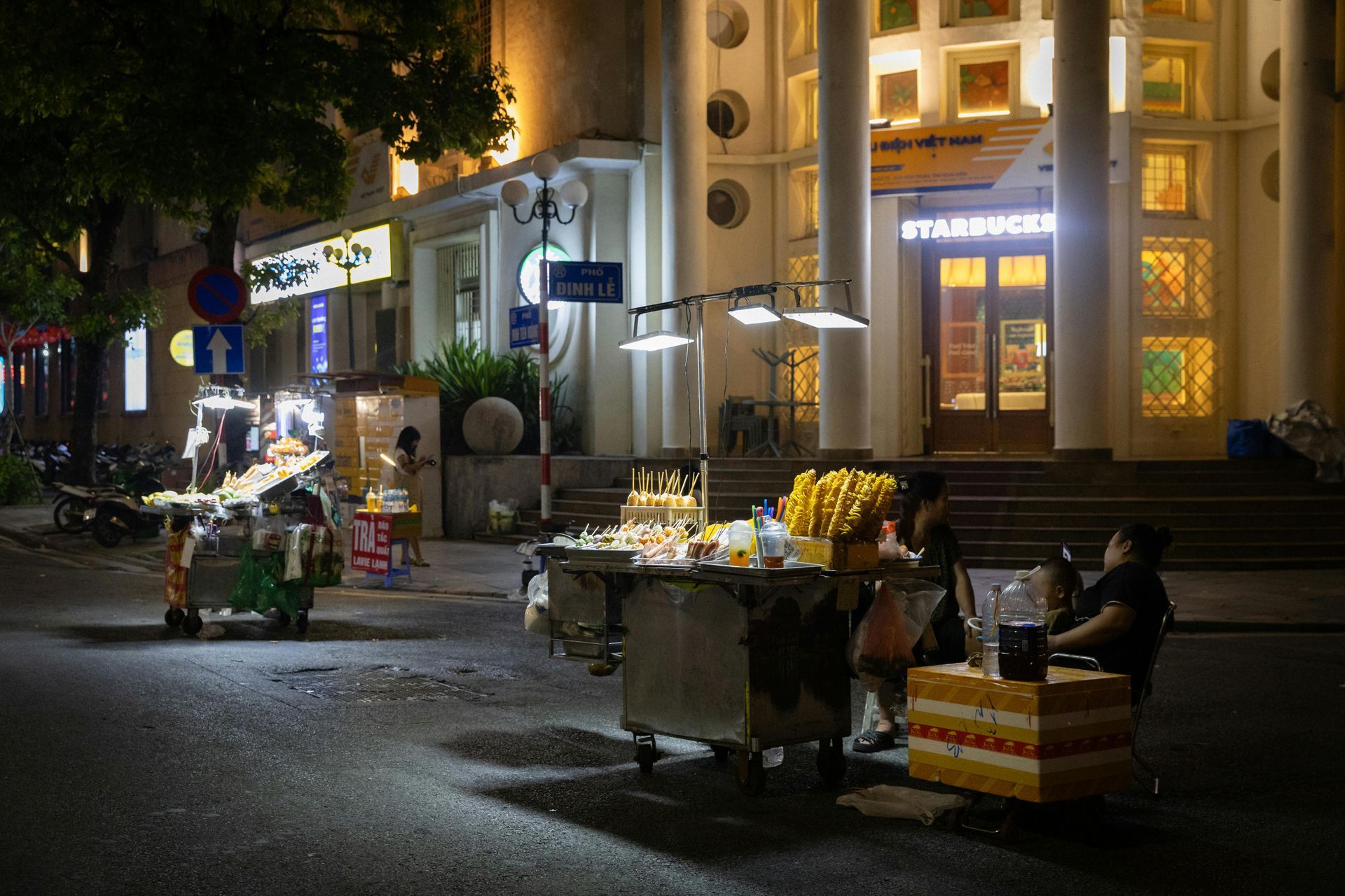 Night street scene with food carts, illuminated Starbucks entrance, and people sitting at a table.