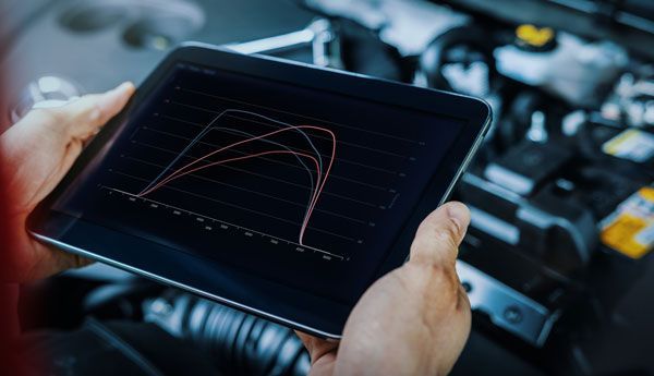 A person is holding a tablet with a graph on it in front of a car engine.