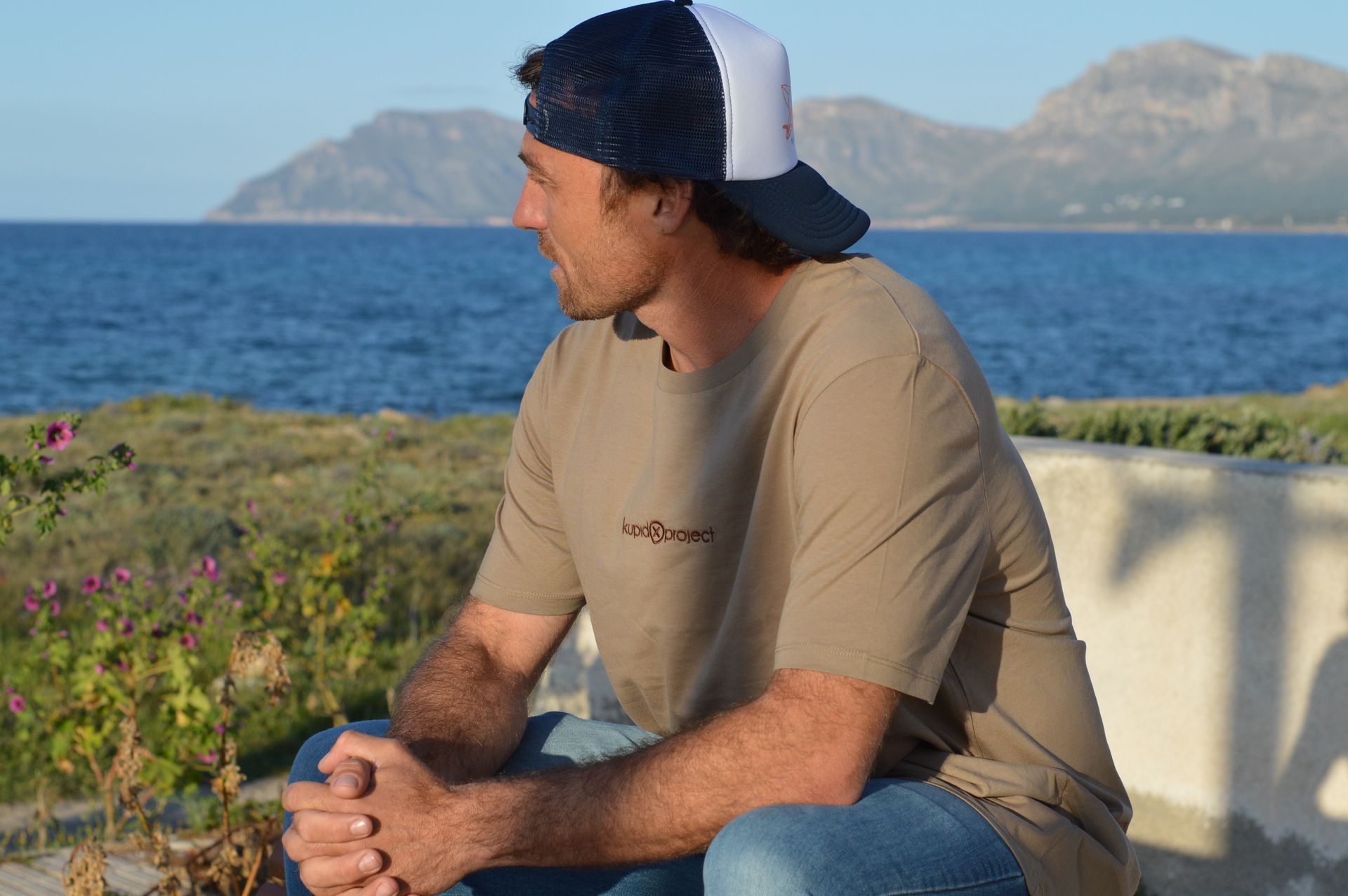 Un hombre con gorra y camiseta Kupido está sentado frente al océano.