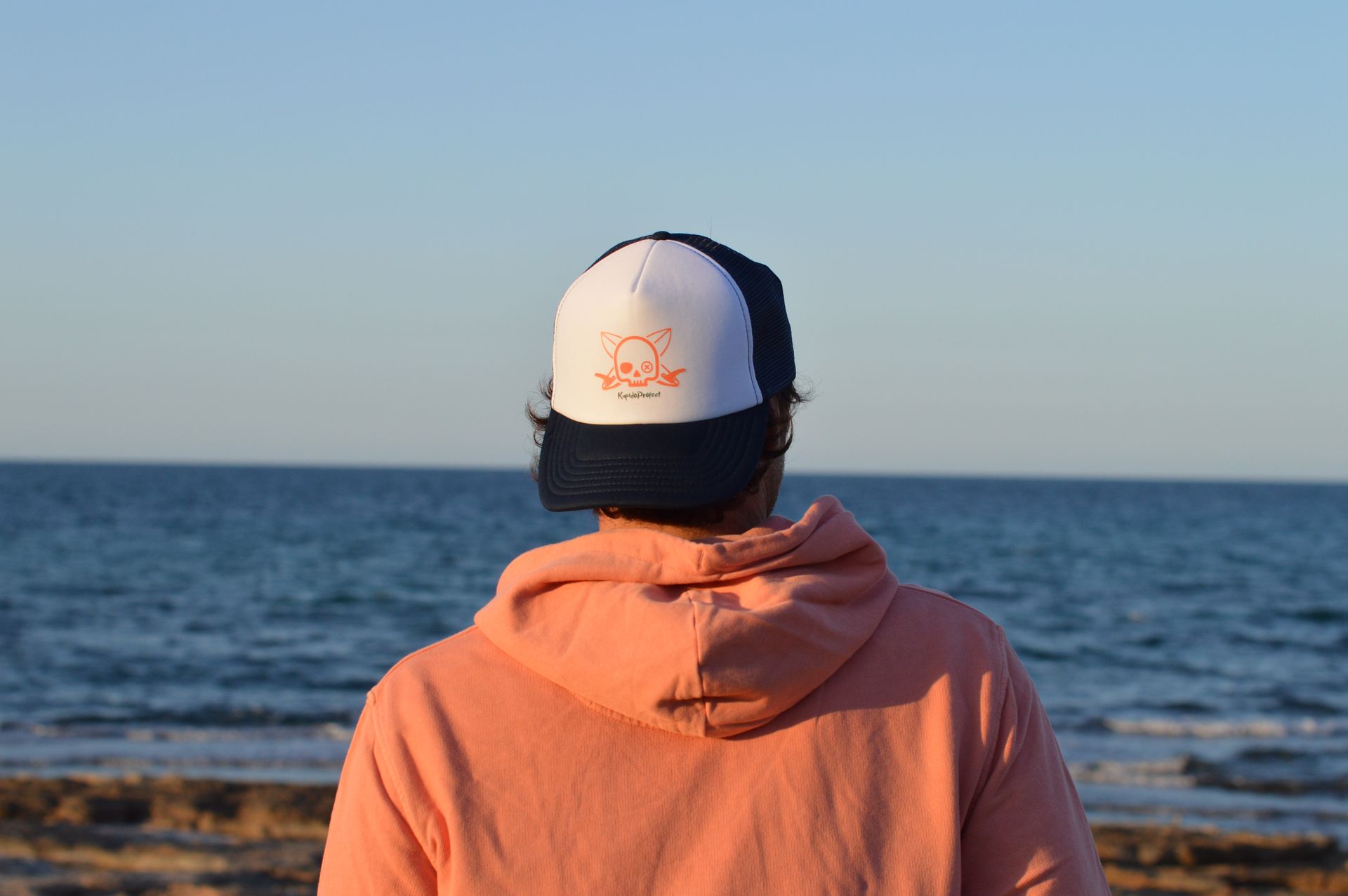 Un hombre con sombrero y sudadera con capucha está parado en una playa mirando el océano.