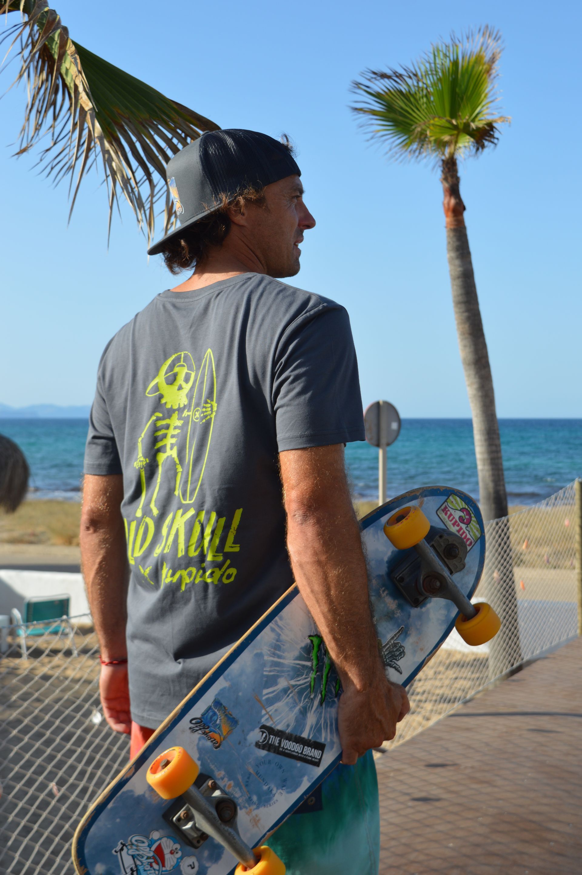 Un hombre sosteniendo una patineta frente a una palmera.
