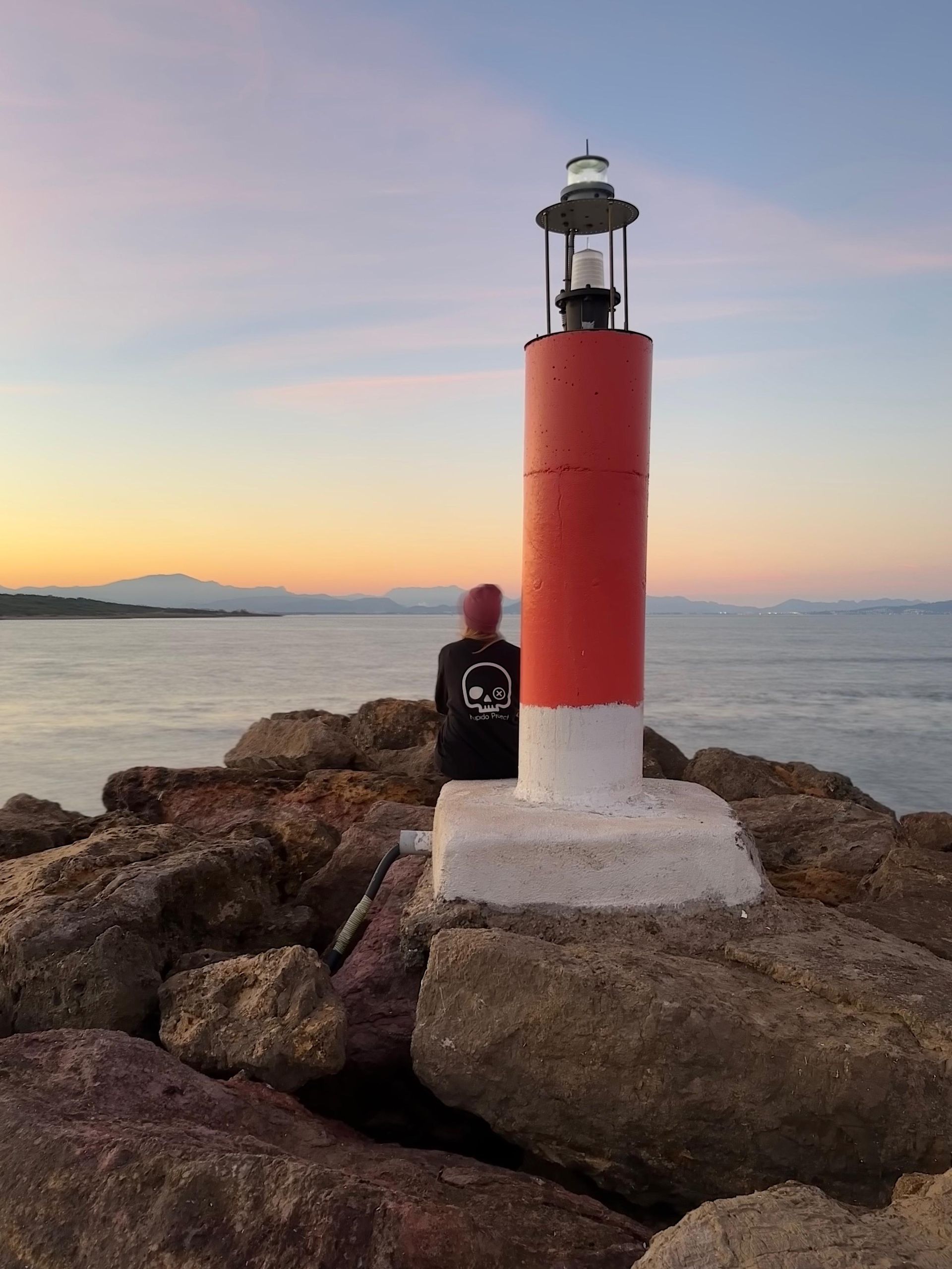 Una persona está sentada junto a un faro rojo y blanco en una costa rocosa.