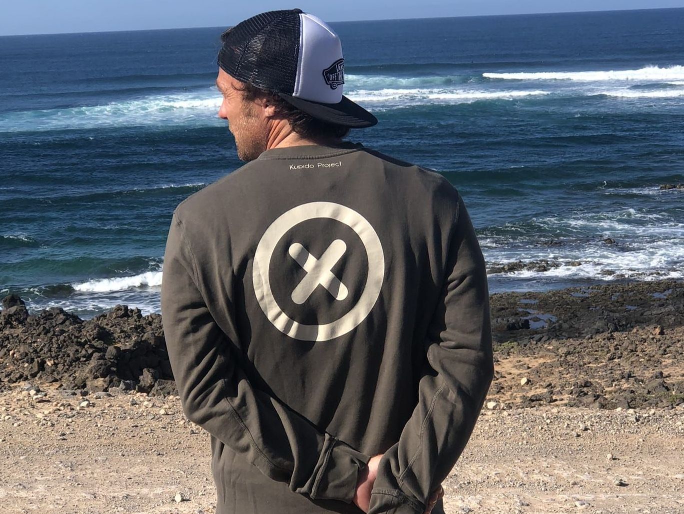 Un hombre con sombrero y sudadera está parado en una playa mirando el océano.