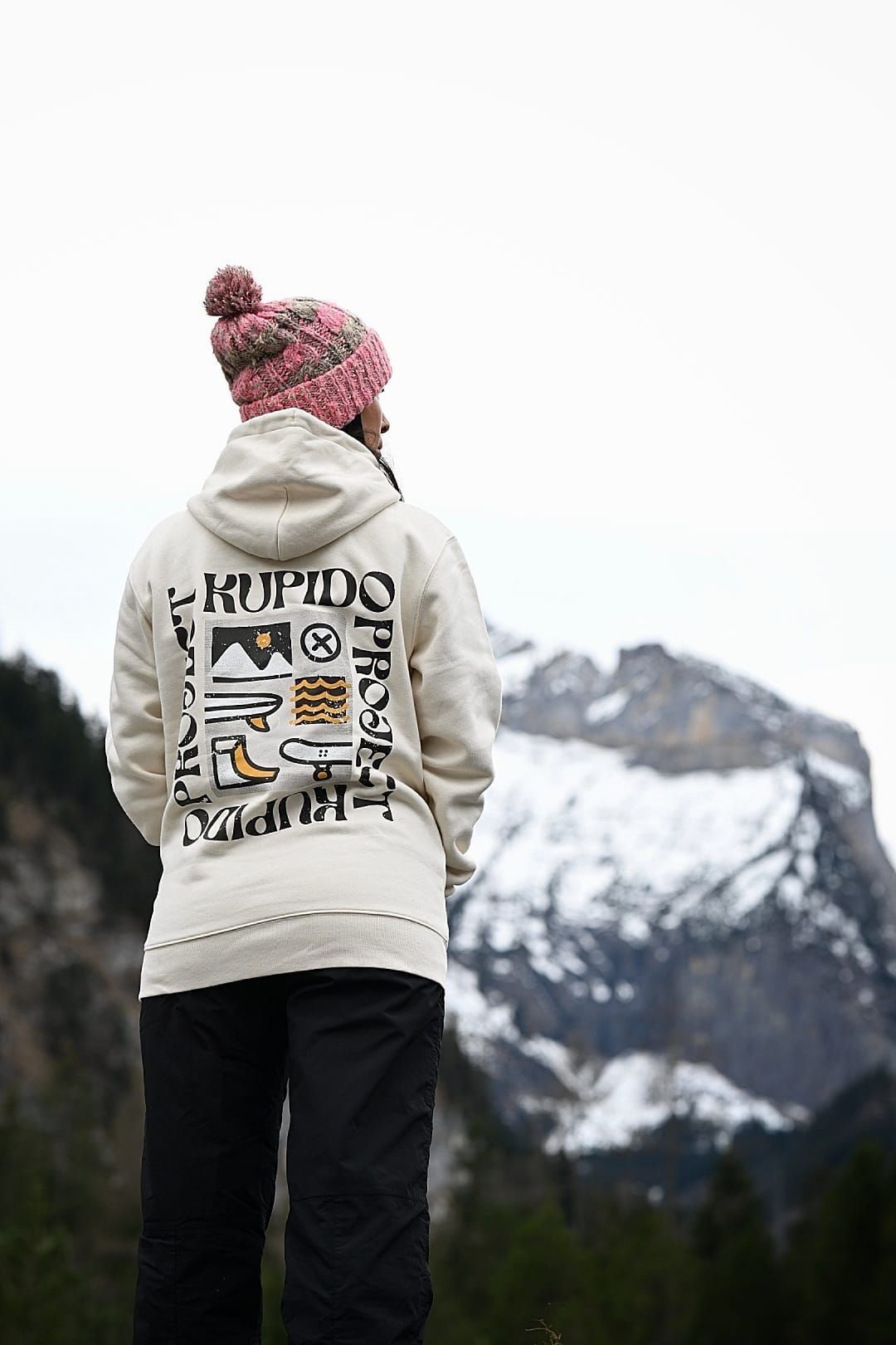 Una persona que lleva una sudadera con capucha y un sombrero está parada frente a una montaña nevada.