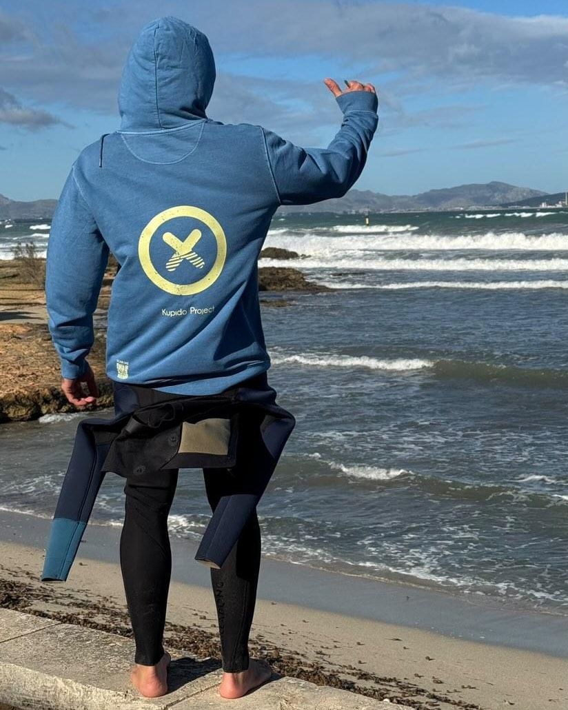 Un hombre con una sudadera con capucha azul está parado en una playa mirando el océano.