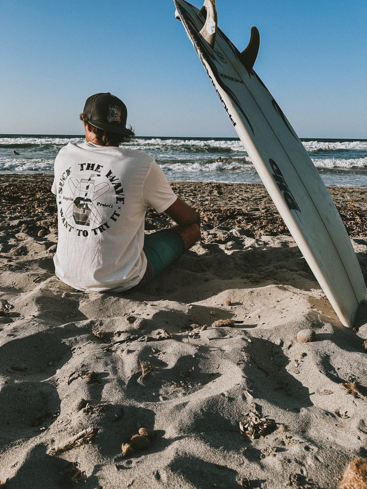 Un hombre está sentado en la playa junto a una tabla de surf.