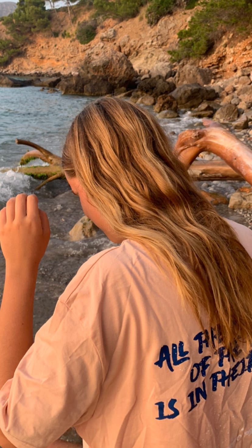 Una chica rubia con una camiseta color melocotón frente al mar.