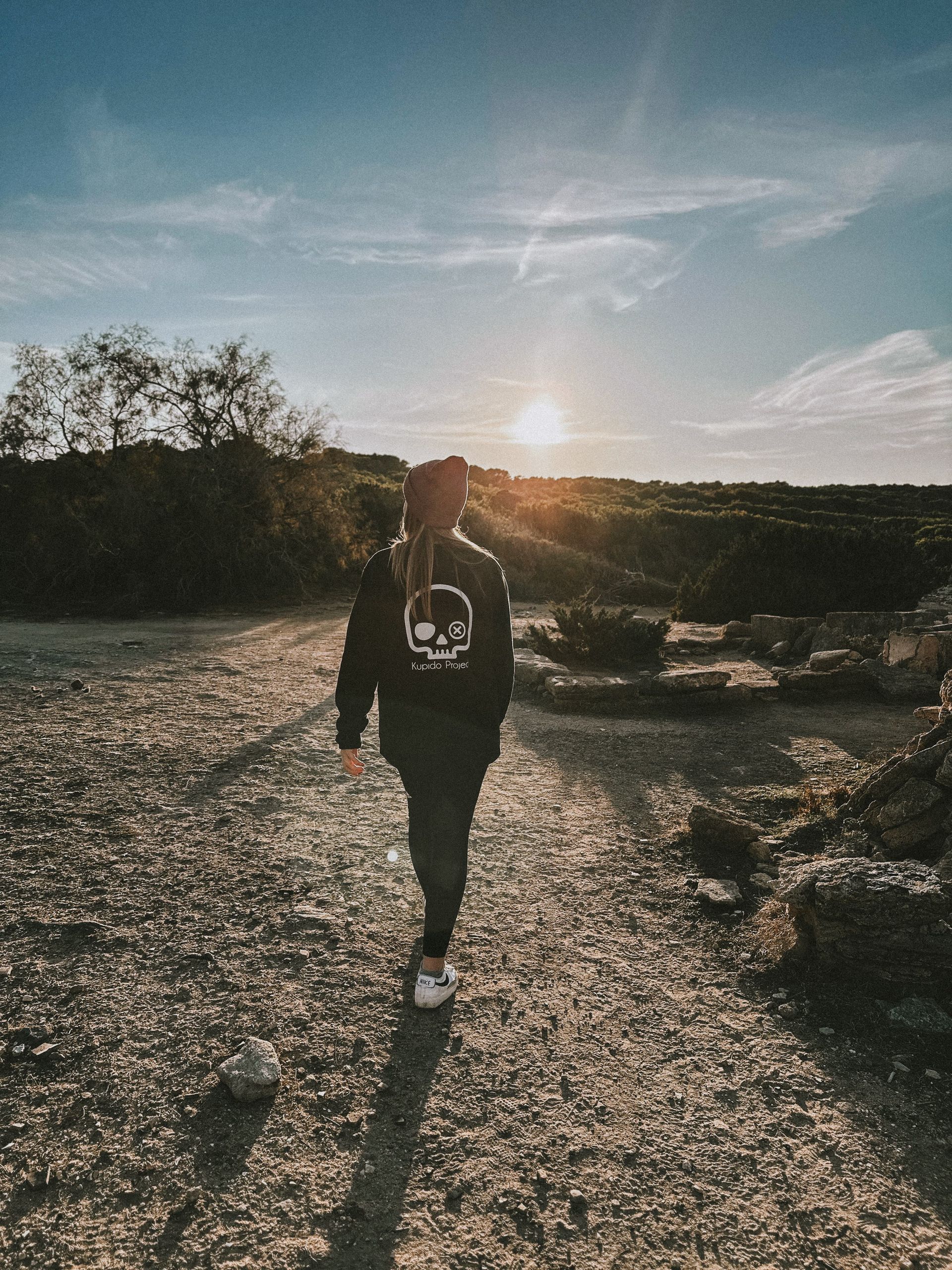 Una persona que viste una sudadera con capucha negra con una calavera camina por un camino de tierra.