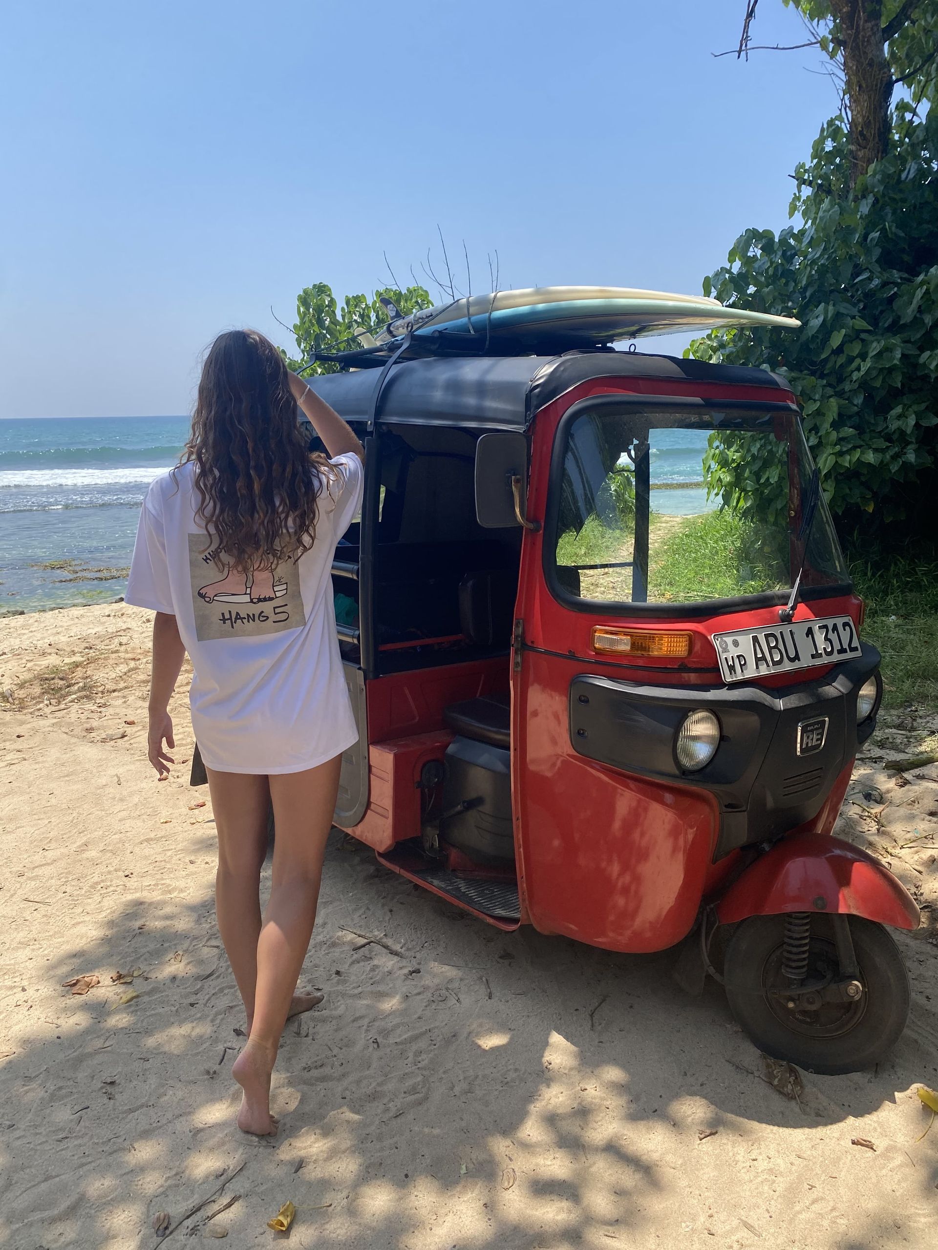 Una mujer camina hacia un rickshaw rojo con una tabla de surf encima.