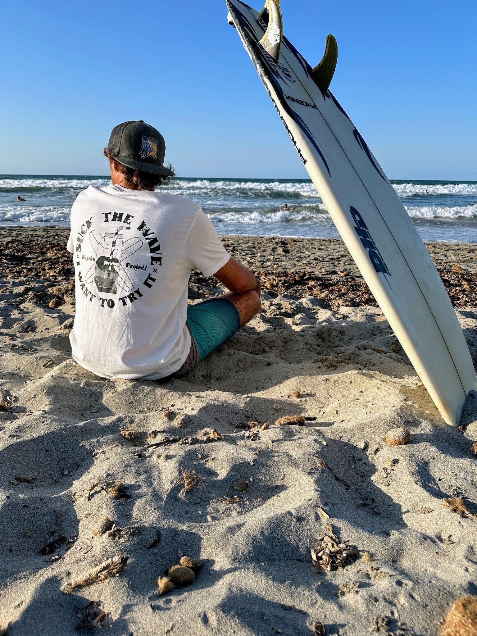 Un hombre está sentado en la playa junto a una tabla de surf.
